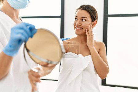 Téléchargez les photos : Jeune femme latine portant une serviette visage regardant sur le miroir au centre de beauté - en image libre de droit