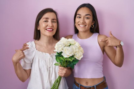 Foto de Madre e hija hispanas sosteniendo ramo de flores blancas mirando confiadas con sonrisa en la cara, señalándose con los dedos orgullosos y felices. - Imagen libre de derechos