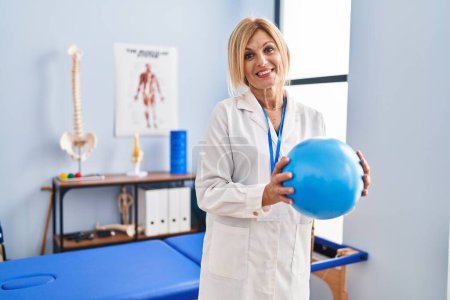 Téléchargez les photos : Middle age blonde woman wearing physiotherapist uniform holding ball at physiotherapy clinic - en image libre de droit