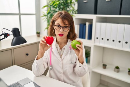 Téléchargez les photos : Young doctor woman holding heart and green apple skeptic and nervous, frowning upset because of problem. negative person. - en image libre de droit
