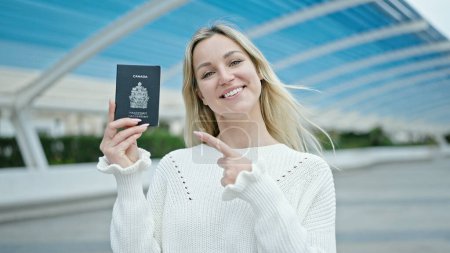 Photo for Young blonde woman smiling confident pointing to canada passport at park - Royalty Free Image