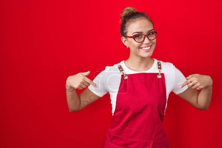 Foto de Mujer hispana joven vistiendo delantal de camarera sobre fondo rojo mirando confiado con sonrisa en la cara, señalándose con los dedos orgullosos y felices. - Imagen libre de derechos