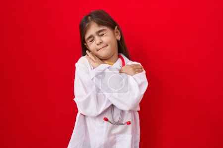 Téléchargez les photos : Petite fille hispanique portant l'uniforme de médecin et stéthoscope se serrant dans ses bras heureux et positifs, souriant confiant. amour de soi et soins de soi - en image libre de droit