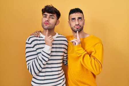 Téléchargez les photos : Jeune couple gay hispanique debout sur fond jaune pensant concentré sur le doute avec le doigt sur le menton et regardant vers le haut se demandant - en image libre de droit