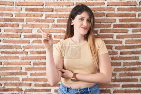 Téléchargez les photos : Jeune femme brune debout sur un mur de briques pointant avec le doigt de la main sur le côté montrant la publicité, visage sérieux et calme - en image libre de droit