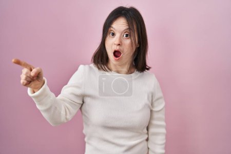 Foto de Mujer con síndrome de Down de pie sobre fondo rosa sorprendida señalando con el dedo hacia un lado, boca abierta expresión asombrada. - Imagen libre de derechos