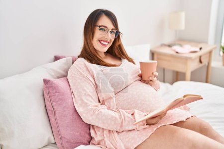Foto de Joven embarazada bebiendo taza de café libro de lectura en el dormitorio - Imagen libre de derechos