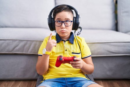 Junge hispanische Kind spielt Videospiel mit Controller trägt Kopfhörer zeigt nach oben Blick traurig und aufgeregt, zeigt die Richtung mit den Fingern, unglücklich und deprimiert. 