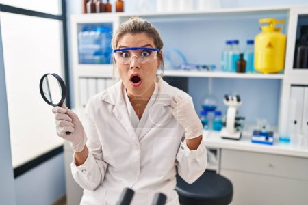 Foto de Mujer joven trabajando en laboratorio científico sosteniendo lupa asustada y sorprendida con la boca abierta para sorpresa, cara de incredulidad - Imagen libre de derechos