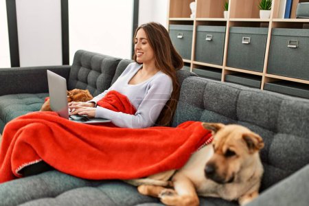 Téléchargez les photos : Jeune femme hispanique utilisant un ordinateur portable assis sur un canapé avec des chiens à la maison - en image libre de droit