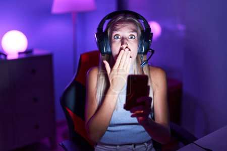 Photo for Young caucasian woman playing video games with smartphone covering mouth with hand, shocked and afraid for mistake. surprised expression - Royalty Free Image