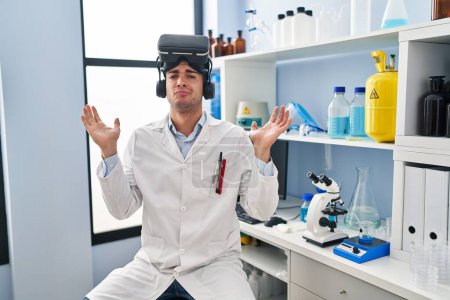 Photo for Young hispanic man working at scientist laboratory wearing vr glasses shouting and screaming loud to side with hand on mouth. communication concept. - Royalty Free Image