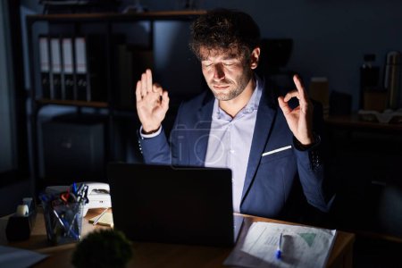 Photo for Hispanic young man working at the office at night relax and smiling with eyes closed doing meditation gesture with fingers. yoga concept. - Royalty Free Image
