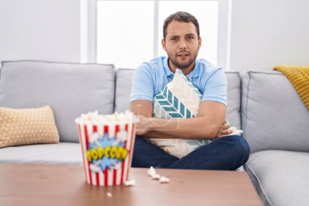 Foto de Hombre hispano viendo televisión en casa sentado en el sofá expresión despistada y confusa. concepto de duda. - Imagen libre de derechos