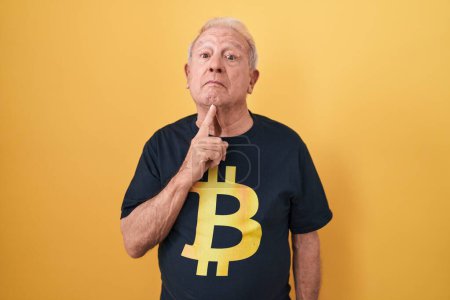 Foto de Hombre mayor con pelo gris usando camiseta bitcoin pensando concentrado en la duda con el dedo en la barbilla y mirando hacia arriba preguntándose - Imagen libre de derechos