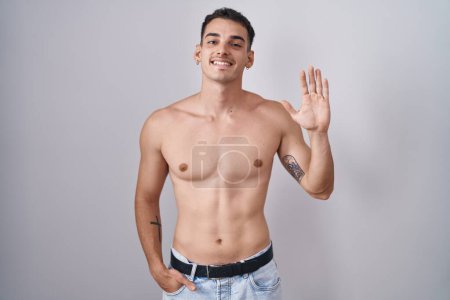 Foto de Hombre hispano guapo de pie sin camisa renunciando a decir hola feliz y sonriente, gesto de bienvenida amistoso - Imagen libre de derechos