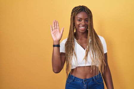 Téléchargez les photos : Femme afro-américaine avec des cheveux tressés debout sur fond jaune renonçant à dire bonjour heureux et souriant, geste de bienvenue amical - en image libre de droit