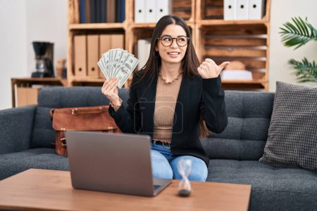 Téléchargez les photos : Jeune femme travaillant au bureau de consultation tenant de l'argent pointant le pouce vers le côté souriant heureux avec la bouche ouverte - en image libre de droit
