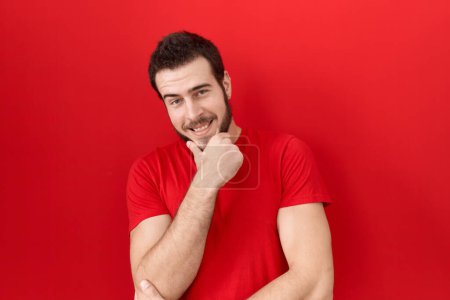 Foto de Joven hombre hispano con camiseta roja casual mirando confiado a la cámara sonriendo con los brazos cruzados y la mano levantada en la barbilla. pensar positivamente. - Imagen libre de derechos