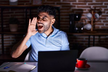 Photo for Hispanic man with beard using laptop at night shouting and screaming loud to side with hand on mouth. communication concept. - Royalty Free Image