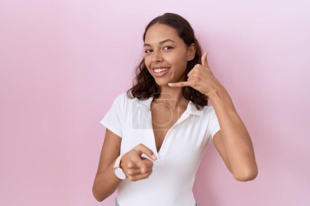 Téléchargez les photos : Jeune femme hispanique portant un t-shirt blanc décontracté souriant faisant parler au téléphone geste et vous pointant du doigt. Appelle-moi. - en image libre de droit