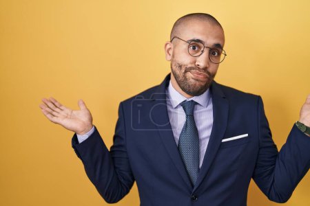 Photo for Hispanic man with beard wearing suit and tie clueless and confused expression with arms and hands raised. doubt concept. - Royalty Free Image