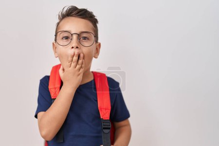 Foto de Pequeño niño hispano con gafas y mochila de estudiante cubriendo la boca con la mano, sorprendido y temeroso de equivocarse. expresión sorprendida - Imagen libre de derechos