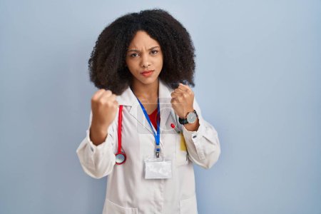 Téléchargez les photos : Jeune femme afro-américaine portant l'uniforme de médecin et stéthoscope prêt à se battre avec geste de défense poing, visage en colère et contrarié, peur du problème - en image libre de droit