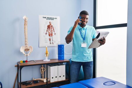 Téléchargez les photos : Young african american man physiotherapist talking on the smartphone al rehab clinic - en image libre de droit