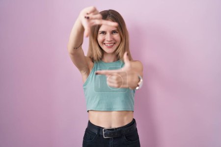 Photo for Blonde caucasian woman standing over pink background smiling making frame with hands and fingers with happy face. creativity and photography concept. - Royalty Free Image