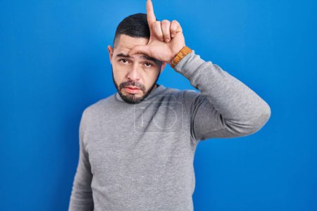 Photo for Hispanic man standing over blue background making fun of people with fingers on forehead doing loser gesture mocking and insulting. - Royalty Free Image