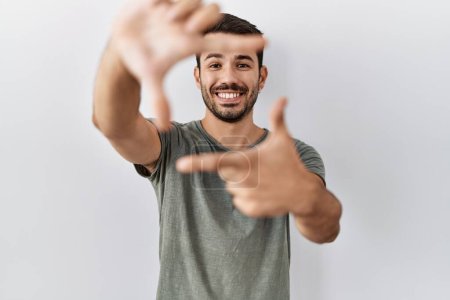 Téléchargez les photos : Jeune homme hispanique avec barbe portant t-shirt décontracté sur fond blanc souriant faisant cadre avec les mains et les doigts avec le visage heureux. concept de créativité et photographie. - en image libre de droit