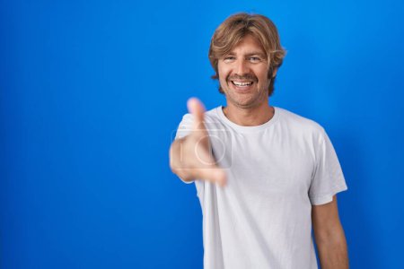 Foto de Hombre de mediana edad de pie sobre fondo azul sonriendo amistoso ofreciendo apretón de manos como saludo y bienvenida. negocios exitosos. - Imagen libre de derechos