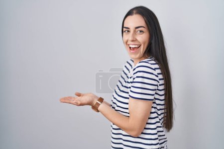 Photo for Young brunette woman wearing striped t shirt pointing aside with hands open palms showing copy space, presenting advertisement smiling excited happy - Royalty Free Image