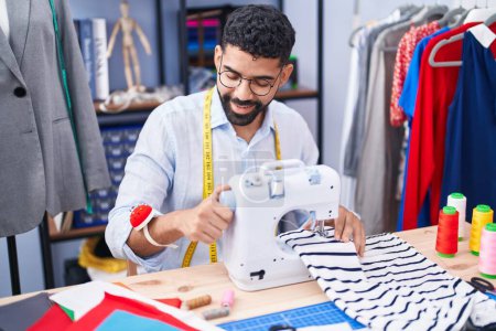 Téléchargez les photos : Jeune tailleur arabe souriant confiant en utilisant une machine à coudre au magasin de tailleur - en image libre de droit