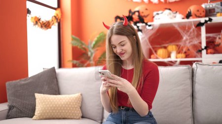 Photo for Young blonde woman smiling wearing devil costume using smartphone at home - Royalty Free Image