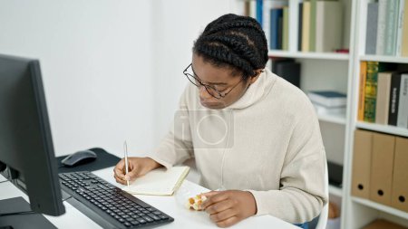 Téléchargez les photos : Femme afro-américaine étudiant écrit sur ordinateur portable manger gaufre à la bibliothèque universitaire - en image libre de droit