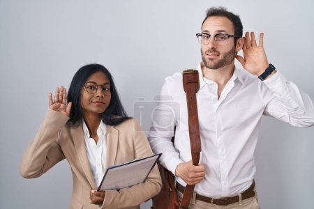 Téléchargez les photos : Couple d'affaires interraciaux portant des lunettes souriant avec la main sur l'oreille à l'écoute d'une audition de rumeurs ou de commérages. concept de surdité. - en image libre de droit