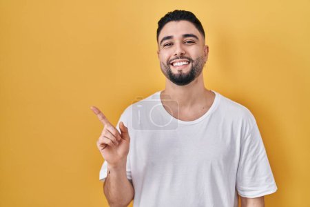 Photo for Young handsome man wearing casual t shirt over yellow background cheerful with a smile of face pointing with hand and finger up to the side with happy and natural expression on face - Royalty Free Image