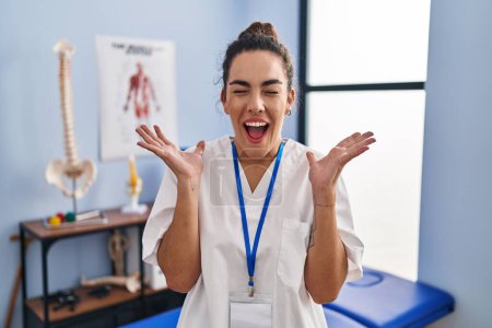 Photo for Young hispanic woman working at rehabilitation clinic celebrating mad and crazy for success with arms raised and closed eyes screaming excited. winner concept - Royalty Free Image