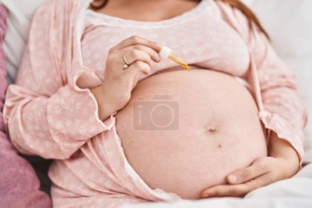 Foto de Mujer embarazada joven aplicando suero al vientre en el dormitorio - Imagen libre de derechos