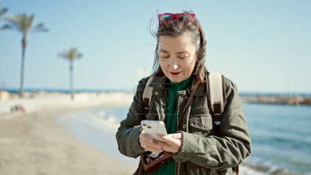 Téléchargez les photos : Femme hispanique mature avec des cheveux gris touriste portant un sac à dos en utilisant un smartphone au bord de la mer - en image libre de droit