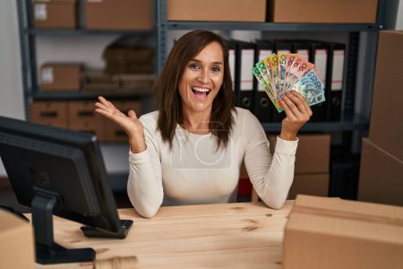 Téléchargez les photos : Femme d'âge moyen travaillant dans un commerce électronique de petite entreprise détenant des dollars australiens célébrant les réalisations avec un sourire heureux et une expression gagnante avec une main levée - en image libre de droit