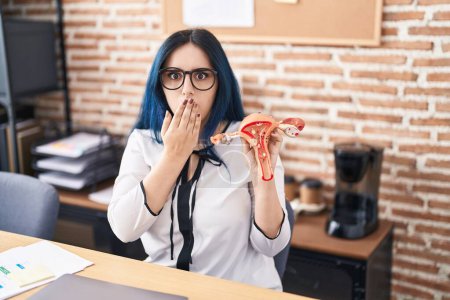 Photo for Young girl with blue hair holding model of female genital organ at the office complaining for menstruation pain covering mouth with hand, shocked and afraid for mistake. surprised expression - Royalty Free Image