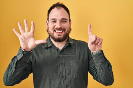 Foto de Hombre hispano de talla grande con barba de pie sobre fondo amarillo mostrando y señalando con los dedos número seis mientras sonríe confiado y feliz. - Imagen libre de derechos