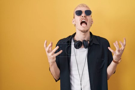 Photo for Young caucasian man wearing sunglasses standing over yellow background crazy and mad shouting and yelling with aggressive expression and arms raised. frustration concept. - Royalty Free Image