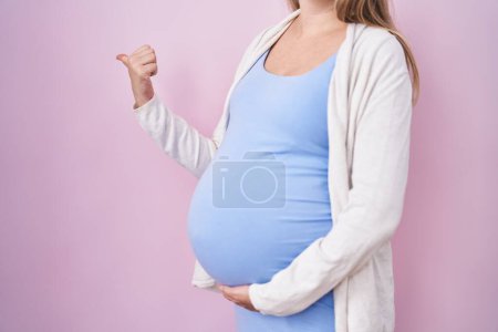 Foto de Mujer embarazada joven esperando un bebé, tocando el vientre embarazada señalando el pulgar hacia el lado sonriendo feliz con la boca abierta - Imagen libre de derechos