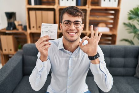 Foto de Joven hombre hispano sosteniendo tarjeta de registro covid haciendo signo ok con los dedos, sonriendo gesto amistoso excelente símbolo - Imagen libre de derechos