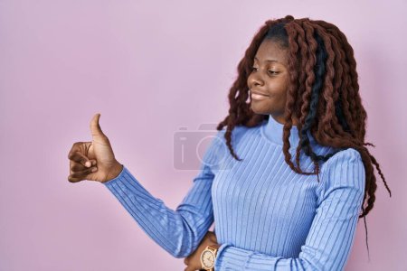 Foto de Mujer africana de pie sobre fondo rosa buscando orgulloso, sonriendo haciendo pulgares hacia arriba gesto a un lado - Imagen libre de derechos