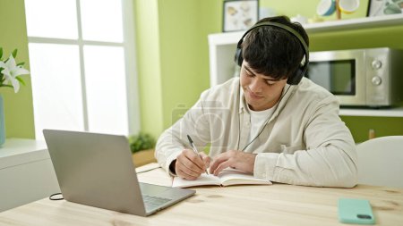 Téléchargez les photos : Jeune homme hispanique étudiant utilisant un ordinateur prenant des notes portant un casque à la salle à manger - en image libre de droit